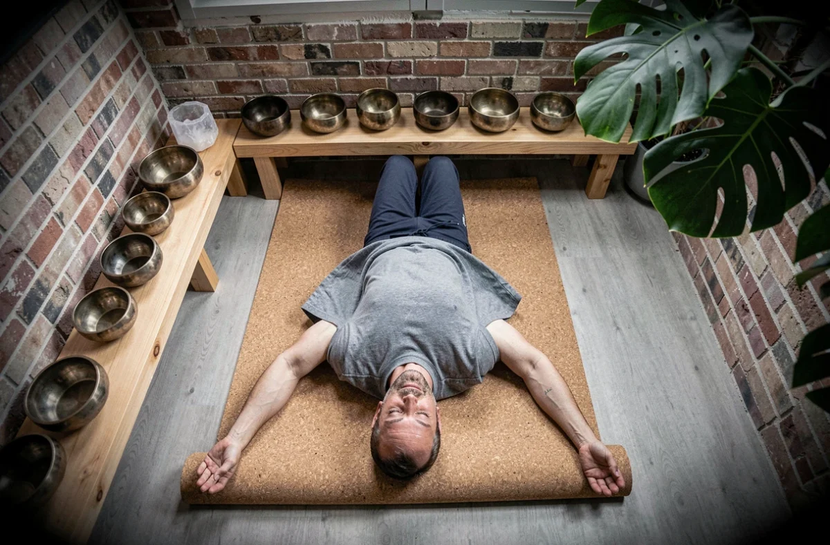 Homme allongé en relaxation profonde sur un tapis en liège entouré de bols chantants dans un salon chaleureux aux murs en briques